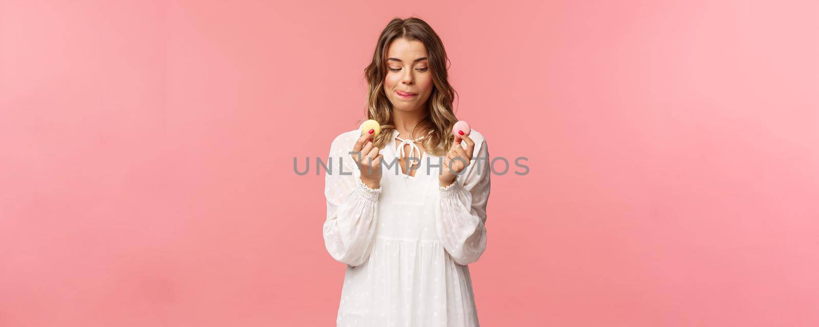 Holidays, spring and party concept. Portrait of cute romantic blond girl in white dress, licking lips as tempting to eat delicious dessert, holding two macarons and look pleased, pink background.