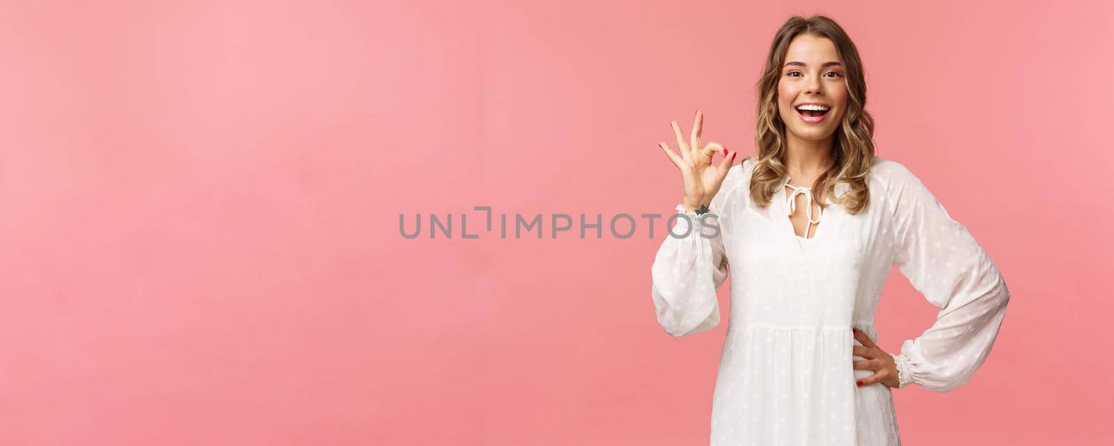 Portrait of carefree good-looking blond girl, wear white dress, guarantee you will like this special spring offer, show okay sign, agree or accept, smiling and nod in approval, pink background.