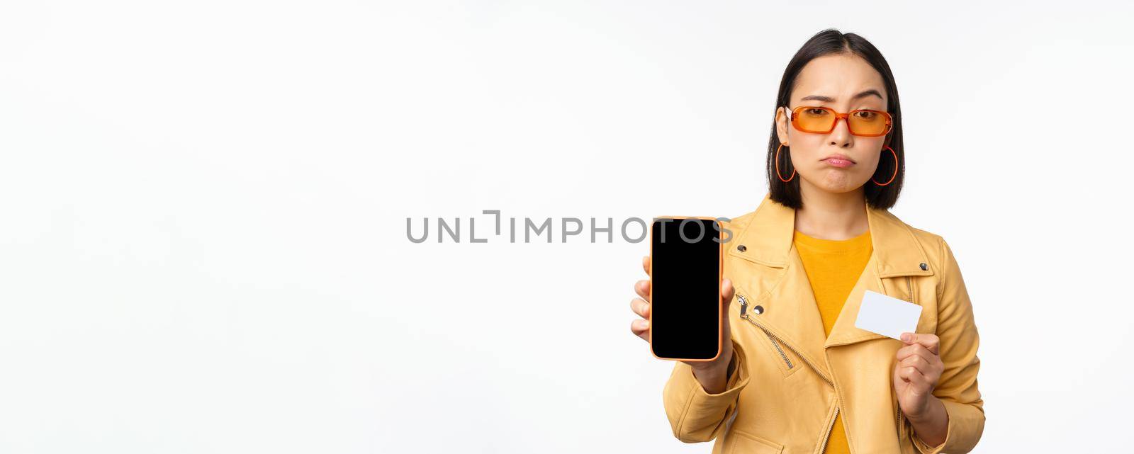 Sad asian girl in sunglasses, showing smartphone app interface, credit card, looking disappointed, standing over white background. Copy space