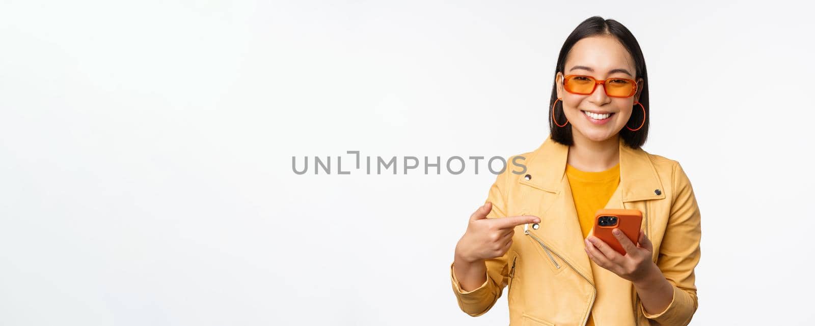 Beautiful smiling asian girl in sunglasses, pointing finger at smartphone, showing app, store on mobile phone, standing over white background.