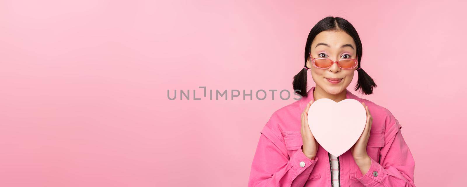 Beautiful asian girl smiling happy, showing heart gift box and looking excited at camera, standing over pink romantic background by Benzoix