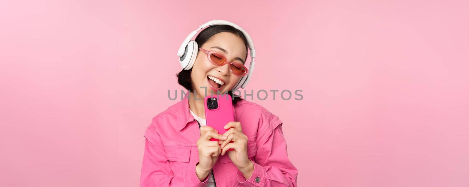 Stylish asian girl in headphones, listening music and taking photos on mobile phone, using smartphone, standing over pink background.