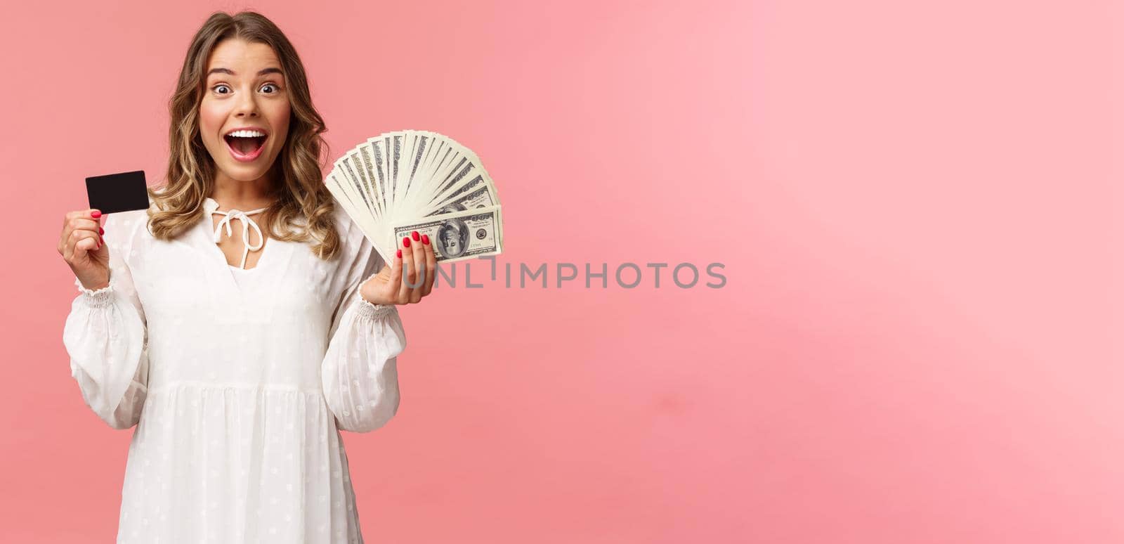 Portrait of excited happy good-looking blond girl in white dress, winning money, placed good bet, made deal, holding dollars money and credit card, smiling amused at camera, pink background by Benzoix