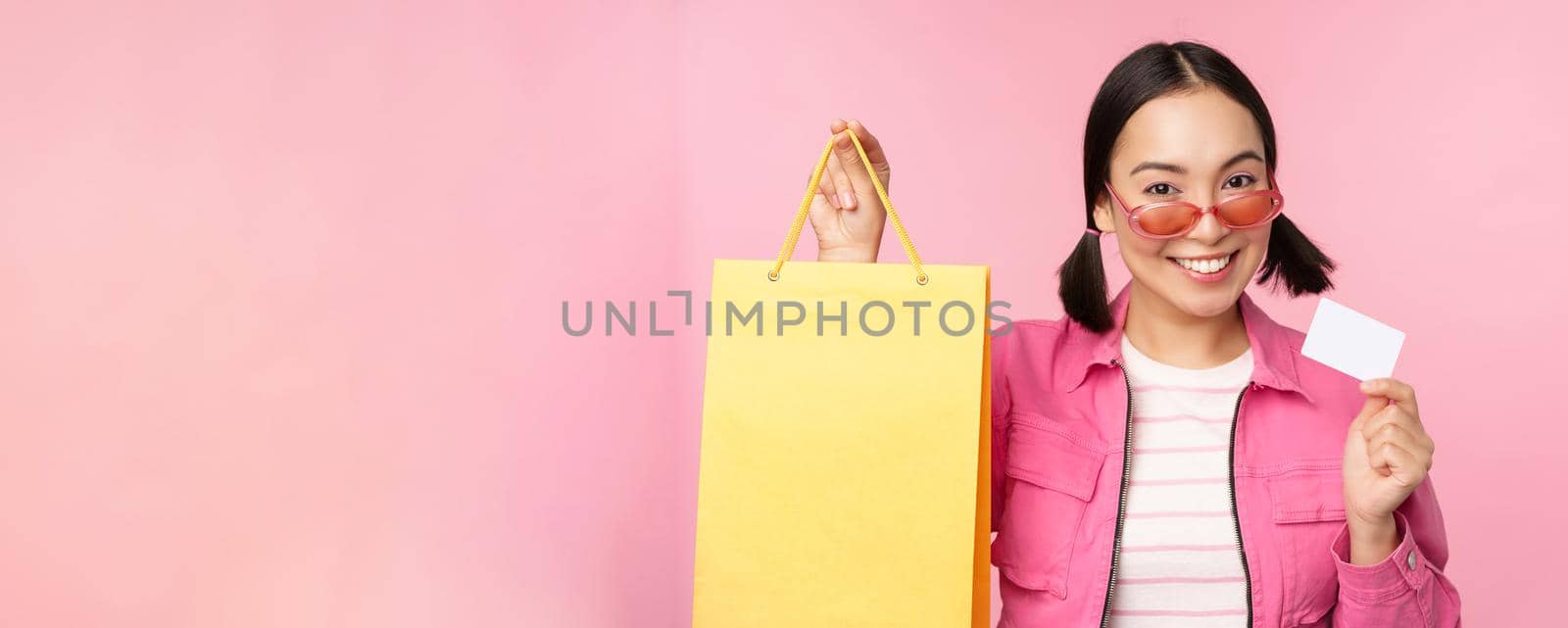 Happy young asian woman shows credit card and shopping bag, store sale announcement, buying smth in shop, posing against pink background.