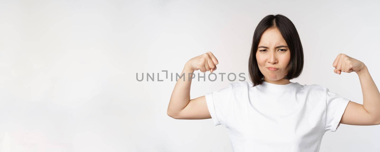 Smiling asian woman showing flexing biceps, muscles strong arms gesture, standing in white tshirt over white background.