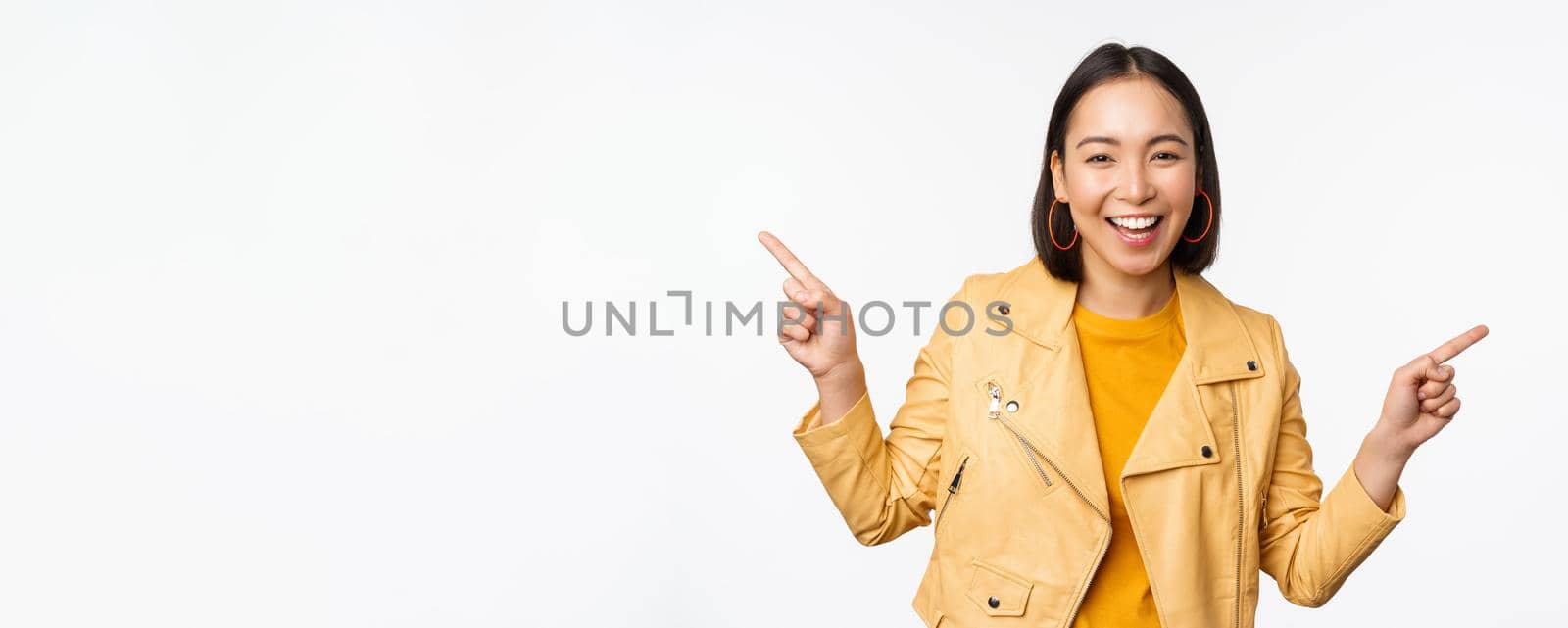 Happy beautiful asian girl pointing fingers left and right, showing banner logo, demonstrating sale, standing in yellow jacket over white background.