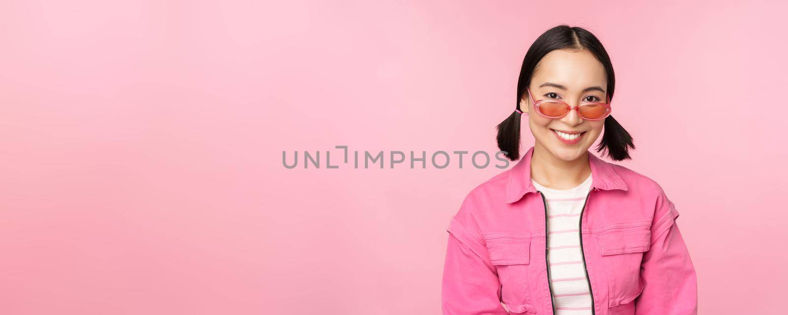 Close up of beautiful asian female model in stylish sunglasses, posing against pink background in trendy outfit, copy space.