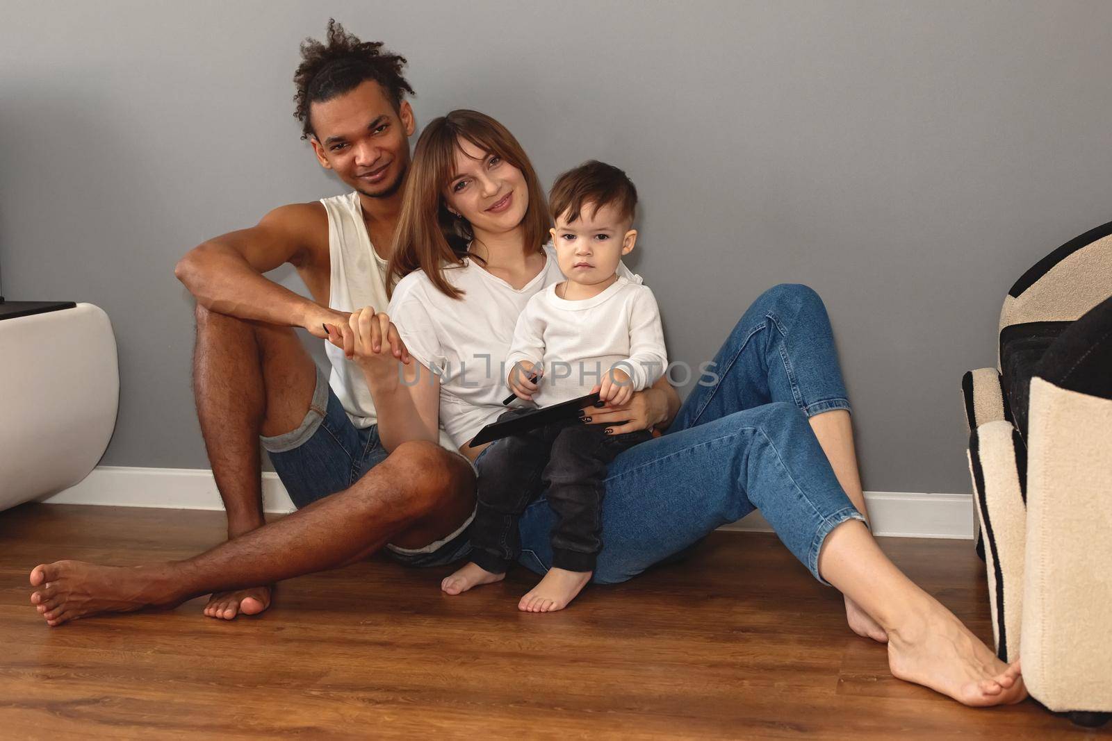 Portrait of happy, modern, multiracial family with a little boy sits against a gray wall in a room by Zakharova