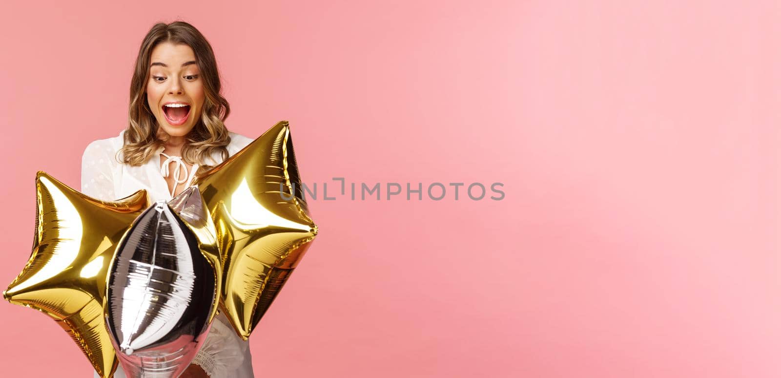 Holidays, celebration and women concept. Portrait of happy lovely young woman in white dress, gasping from amazement and joy, holding birthday star-shaped balloons, pink background.