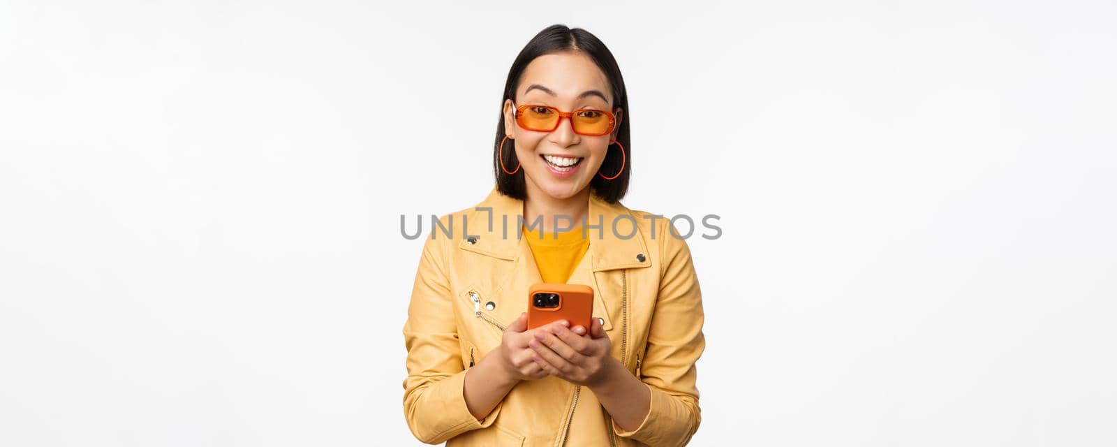 Modern asian girl in sunglasses using her mobile phone, smiling and looking happy, posing against white background. Copy space