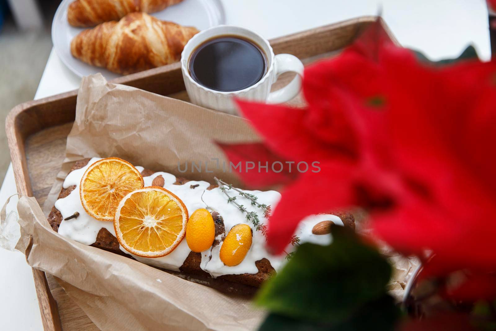 Freshly baked christmas cake and croissant on a gray round plate, a white cup of coffee and a garland on a tray on the table. Blooming poinsettia.