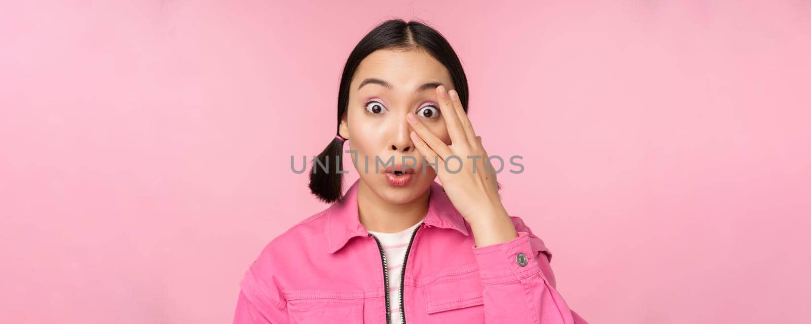 Close up portrait of young asian girl looking surprised, express amazement and wonder, peeking through fingers, standing over pink background.