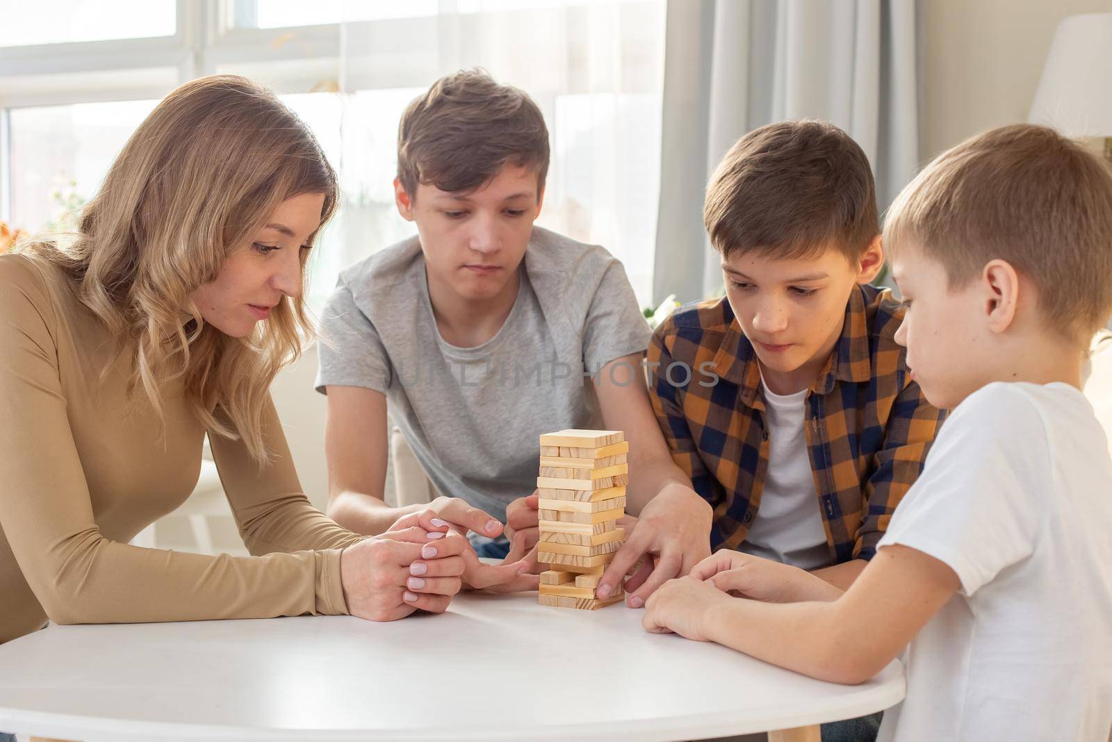 A family, three boys and a woman, are enthusiastically playing a board game made of wooden rectangular blocks stacked in the form of a tower in a sunny room. Close-up