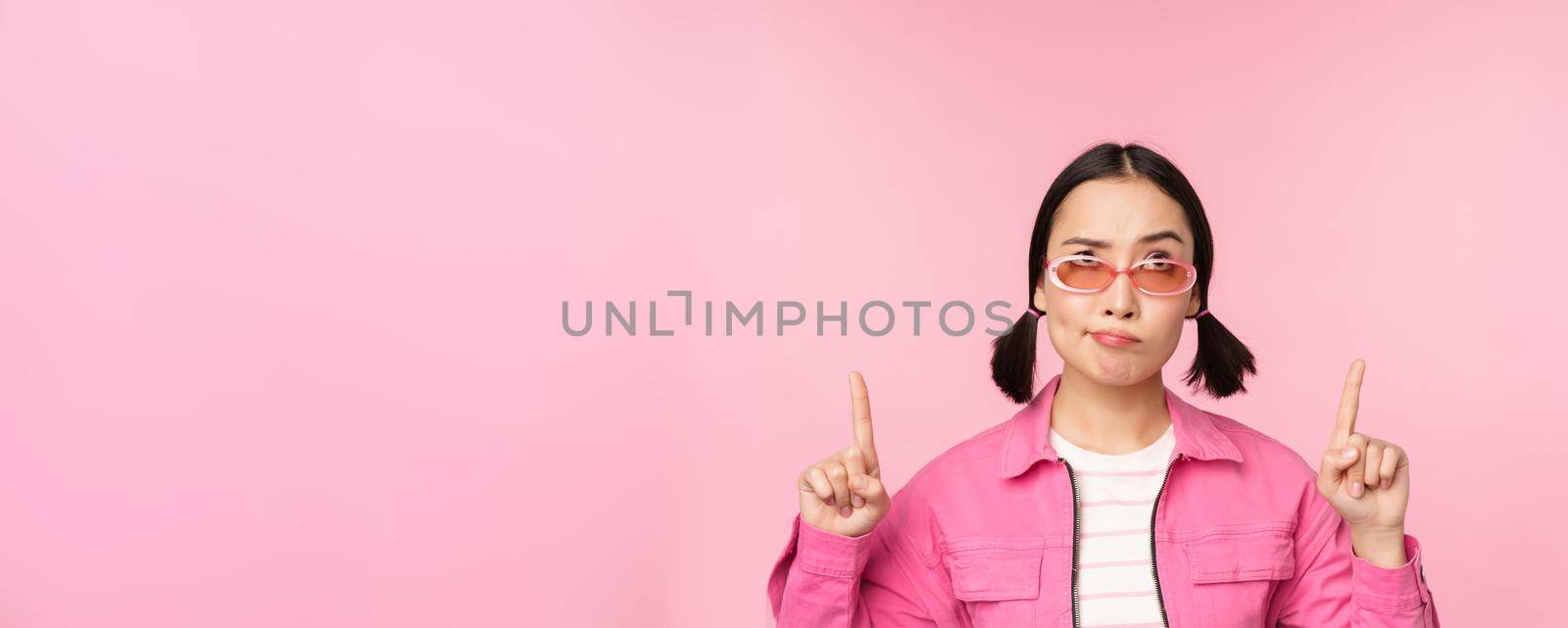 Hesitant asian girl, wears sunglasses, frowns and points fingers up, looks with disbelief, skeptical face expression, stands over pink background.
