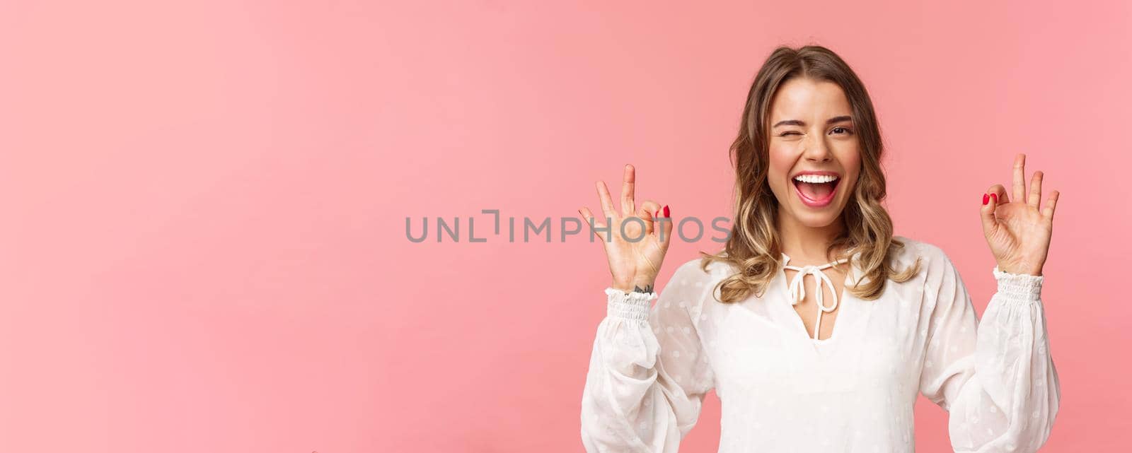 Close-up portrait of beautiful blond girl in white dress guarantee you will enjoy this event, show okay sign, confirm everything excellent, wink and smiling carefree, standing pink background.