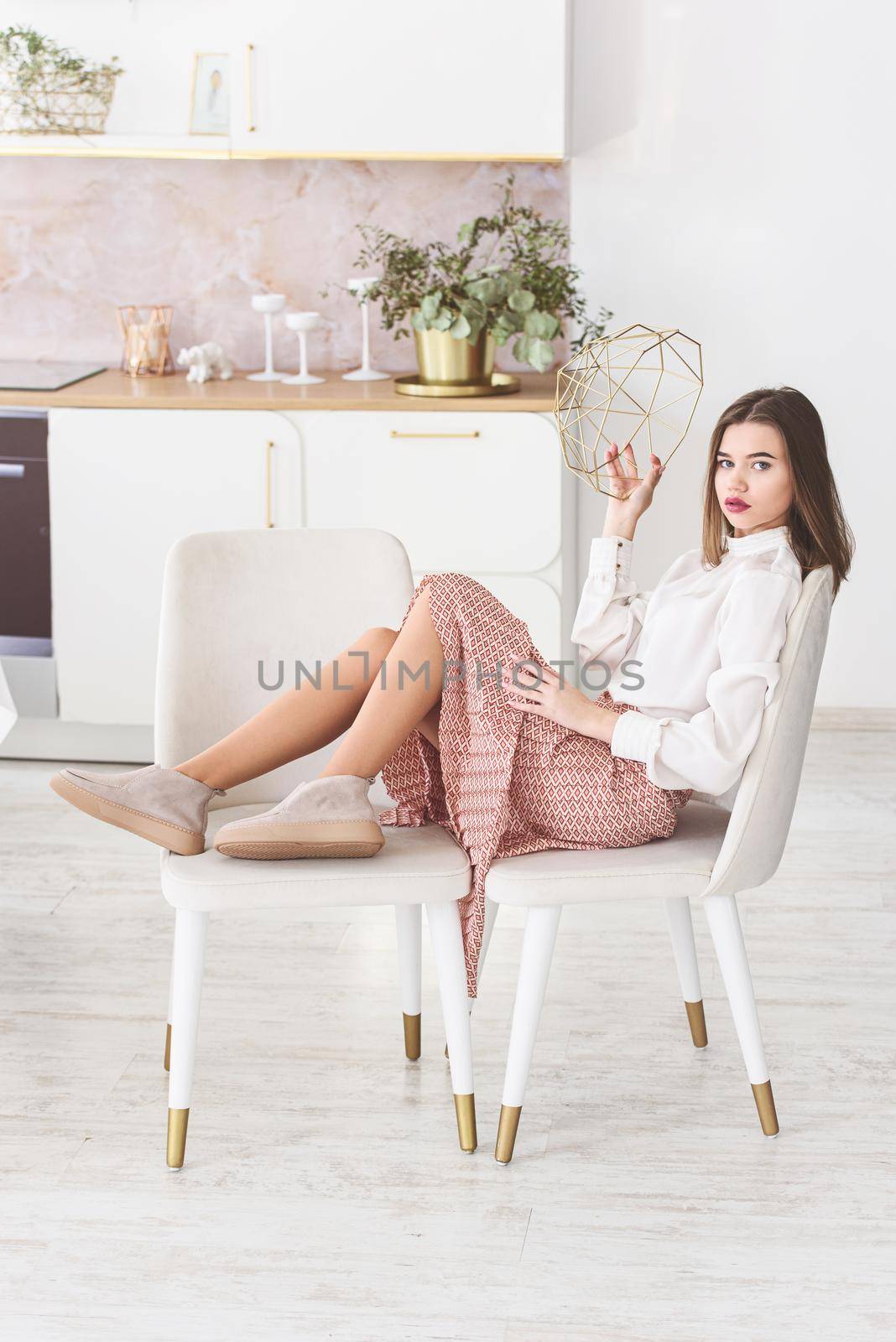 Portrait of fashionable women in beige skirt, white blouse and stylish suede loafer posing on the kitchen in a chair by Ashtray25