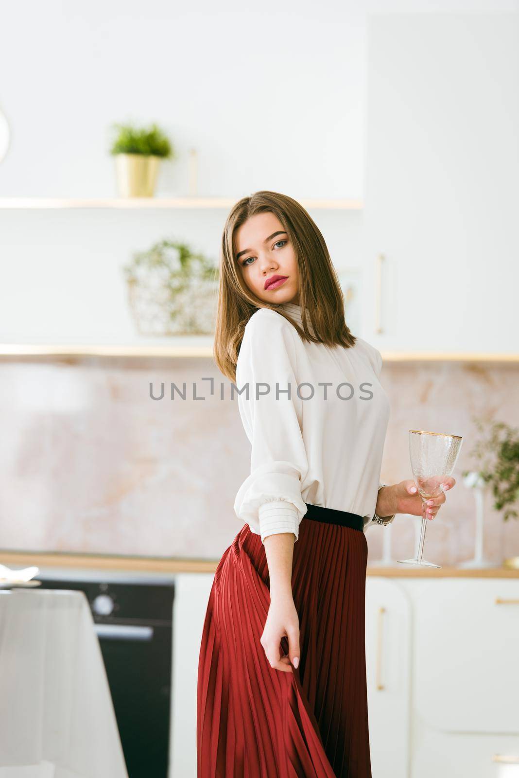 Portrait of fashionable woman in a red skirt and white blouse posing on the kitchen. Girl with a big red lips hold vine glass in a hands