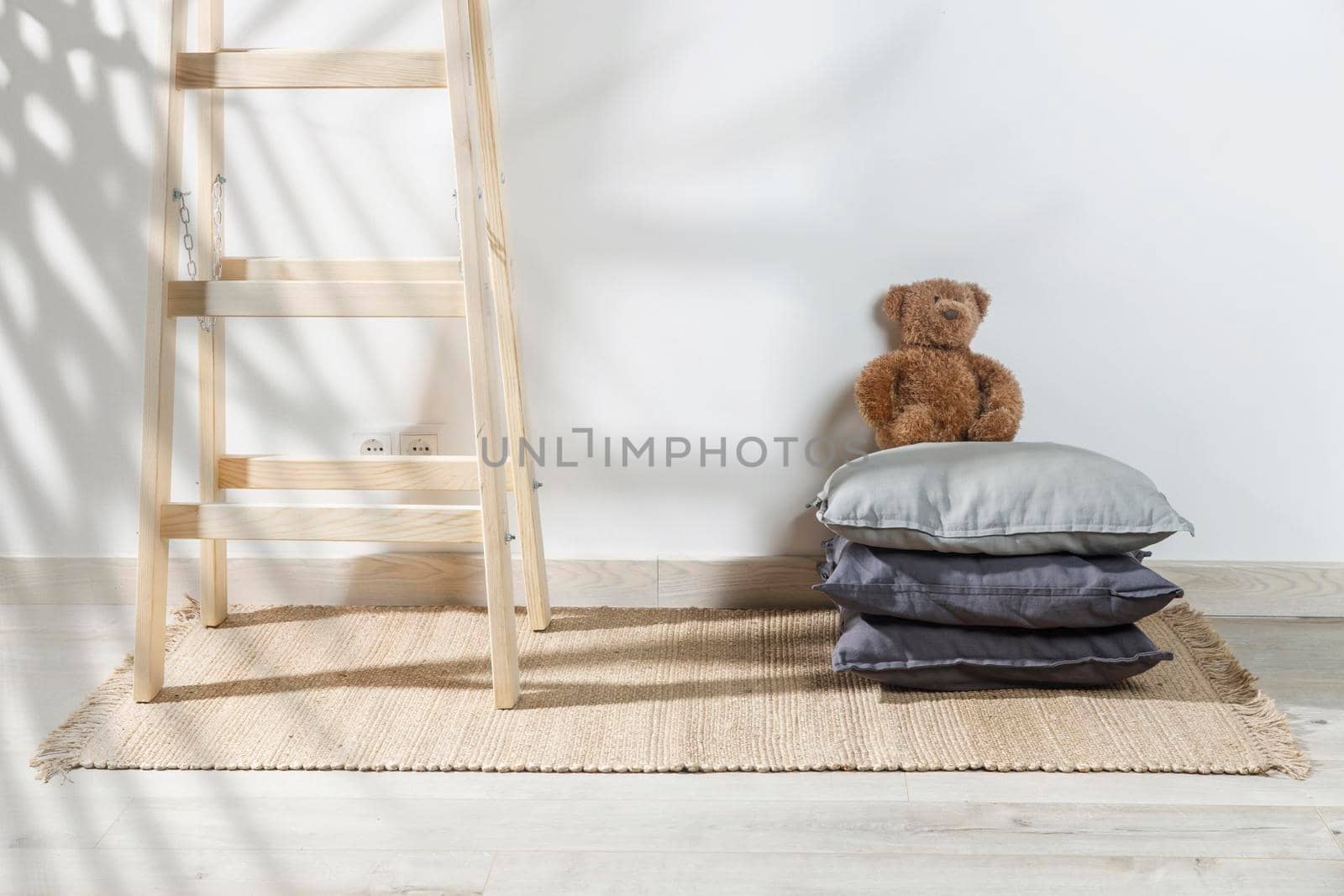 The teddy bear is leaning against the wall next to a small children's stool with three gray pillows on it. The stepladder is leaning against the wall. Copy space