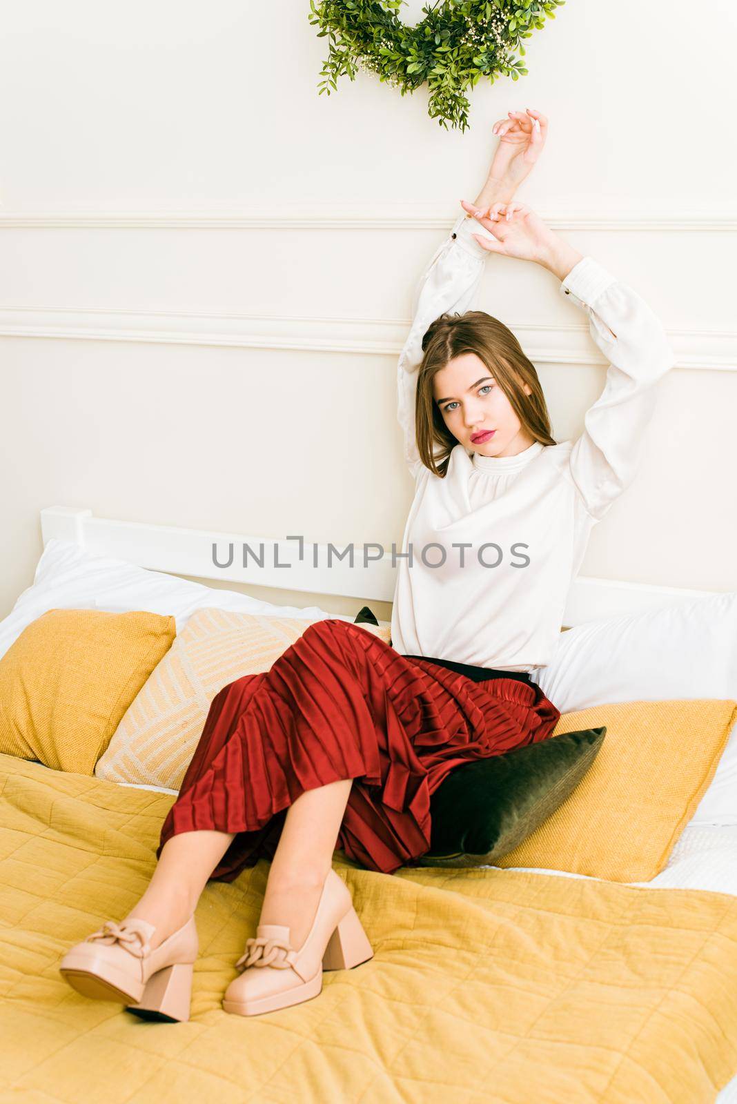 Portrait of fashionable women in red skirt, white blouse and stylish beige high-heeled shoes with a chain buckle posing in a bed. Girl with a big red lips
