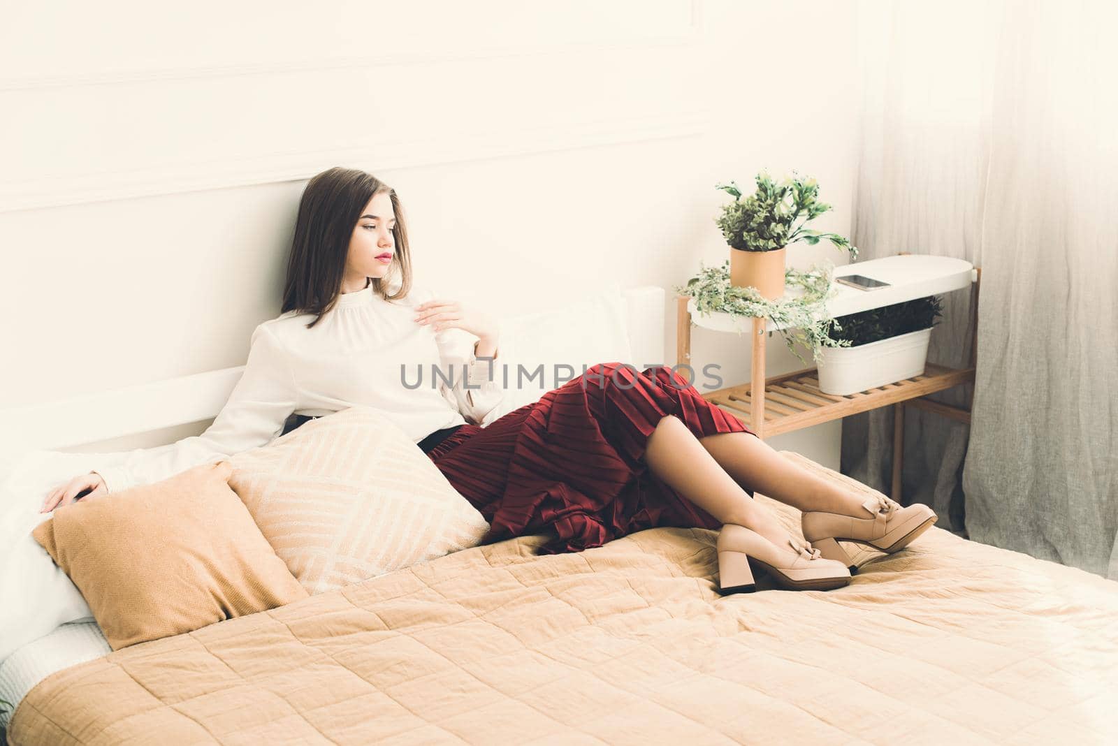 Portrait of fashionable women in red skirt, white blouse and stylish beige high-heeled shoes with a chain buckle posing in a bed. Girl with a big red lips