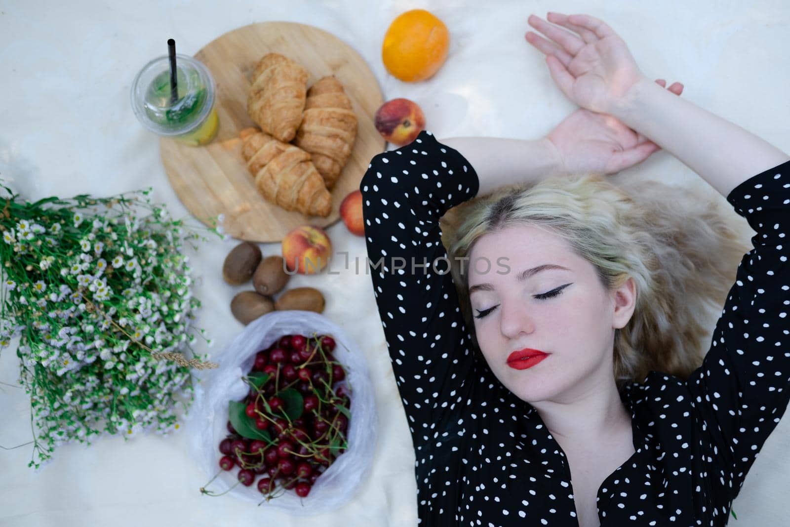 charming blonde young woman on a picnic on plaid in park with tasty snacks. Lemonade, fruits and croissants. summertime, rest, relax, enjoy. freedom.