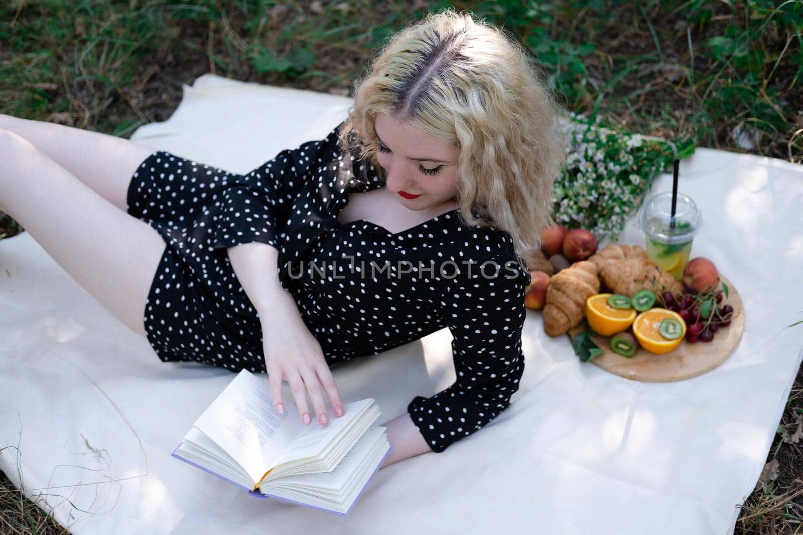 portrait of young woman on a picnic on plaid in park reading a book with tasty snacks.