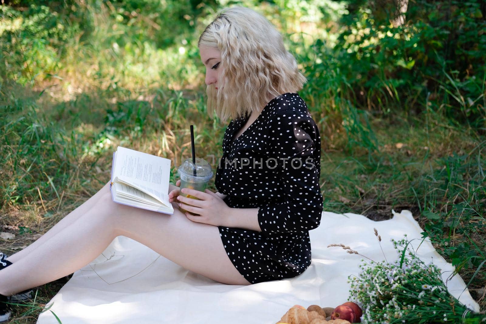 portrait of young woman on a picnic on plaid in park reading a book with tasty snacks.