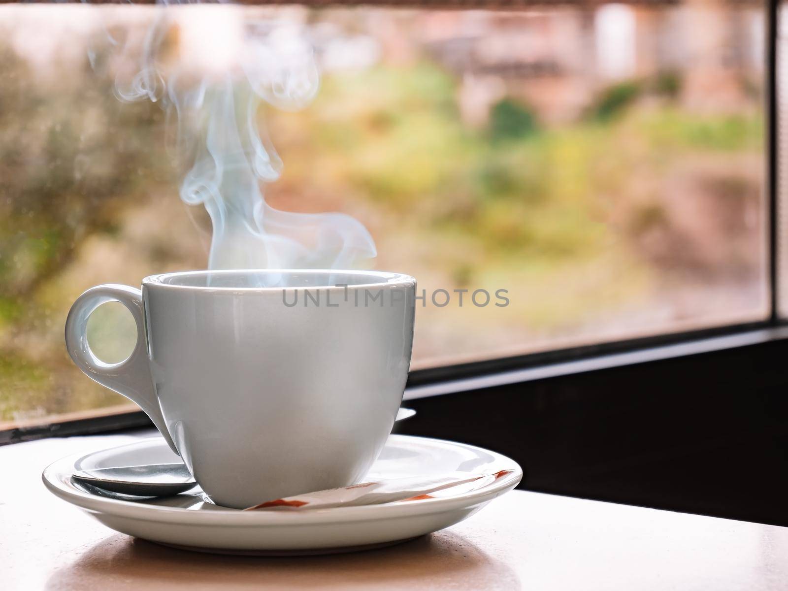 Close up of a white coffee cup on a bar table with a window in the background. Steaming cup of coffee. by CatPhotography