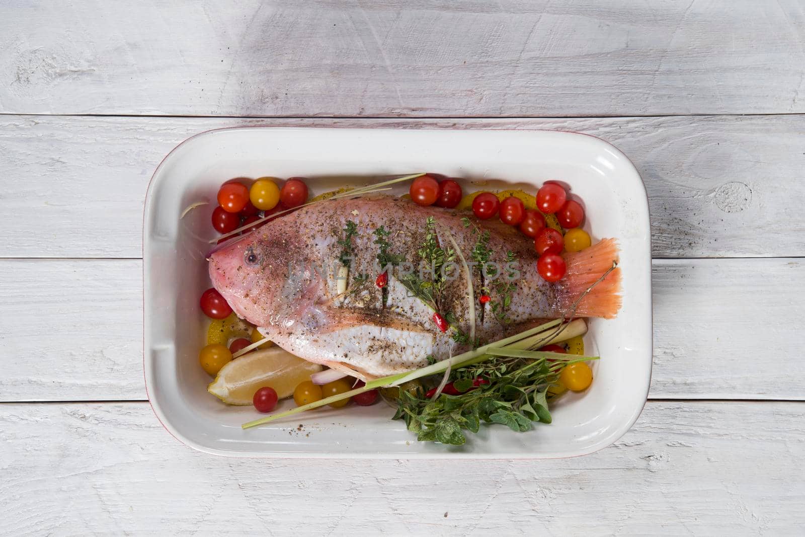 raw pink tilapia fish with vegetables in a baking dish, hands spice on the fish, step by step recipe for white wooden planks,High quality photo