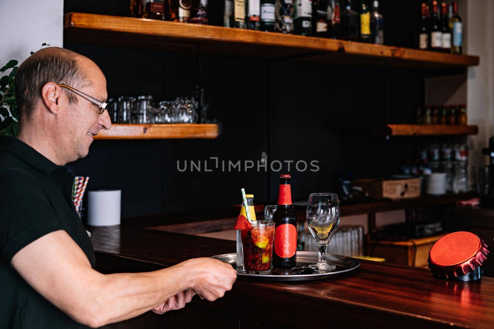 Male waiter, mature, bald, smiling, dressed in company uniform, black polo shirt, Mature, experienced waiter taking an order at the bar counter. worker in his small busines. Worker in his small business. Warm atmosphere and dim lighting. Horizontal