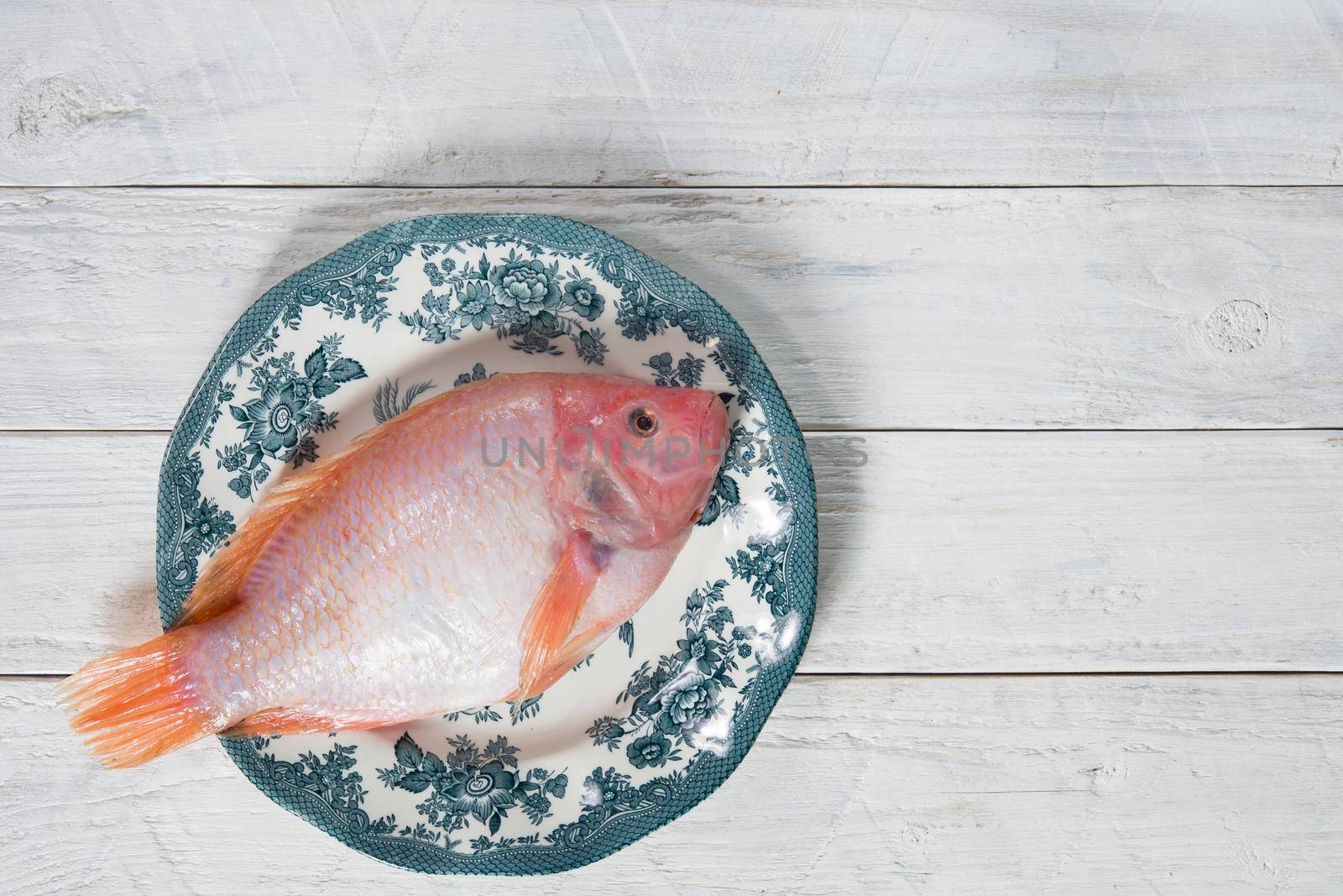 raw pink tilapia fish lies on a plate with blue ornaments on white wooden boards by KaterinaDalemans