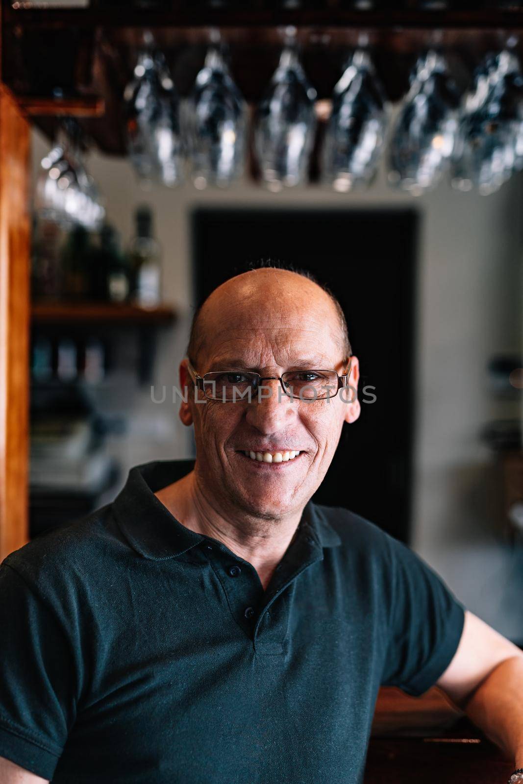 Portrait of an adult male waiter, bald with glasses, smiling, experienced, concentrated and hard-working, dressed in company uniform, a black polo shirt, standing in his small shop. Looking at camera. Warm atmosphere and dim lighting. vertical. glasses hanging in the background over the counter.