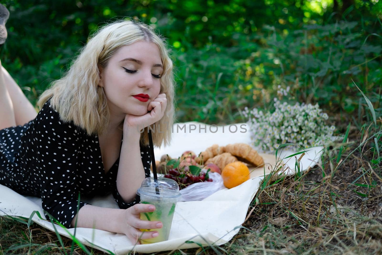 charming blonde young woman on a picnic on plaid in park with tasty snacks. Lemonade, fruits and croissants. summertime, rest, relax, enjoy. freedom.