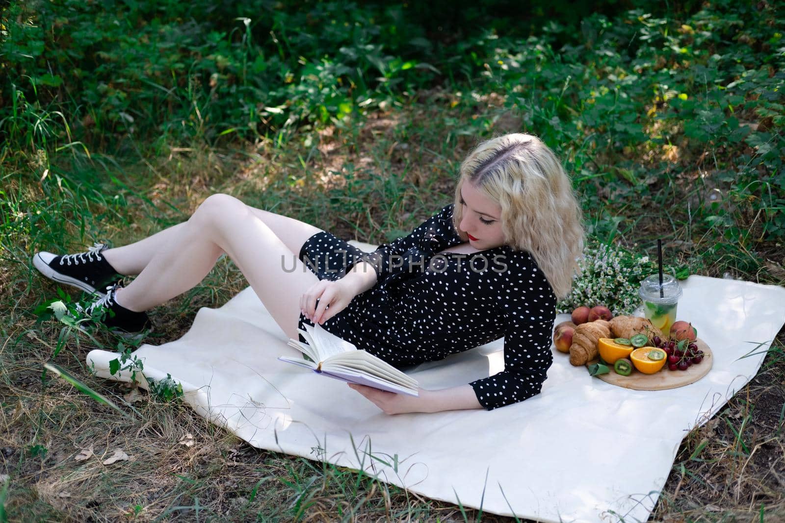 portrait of young woman on a picnic on plaid in park reading a book with tasty snacks.