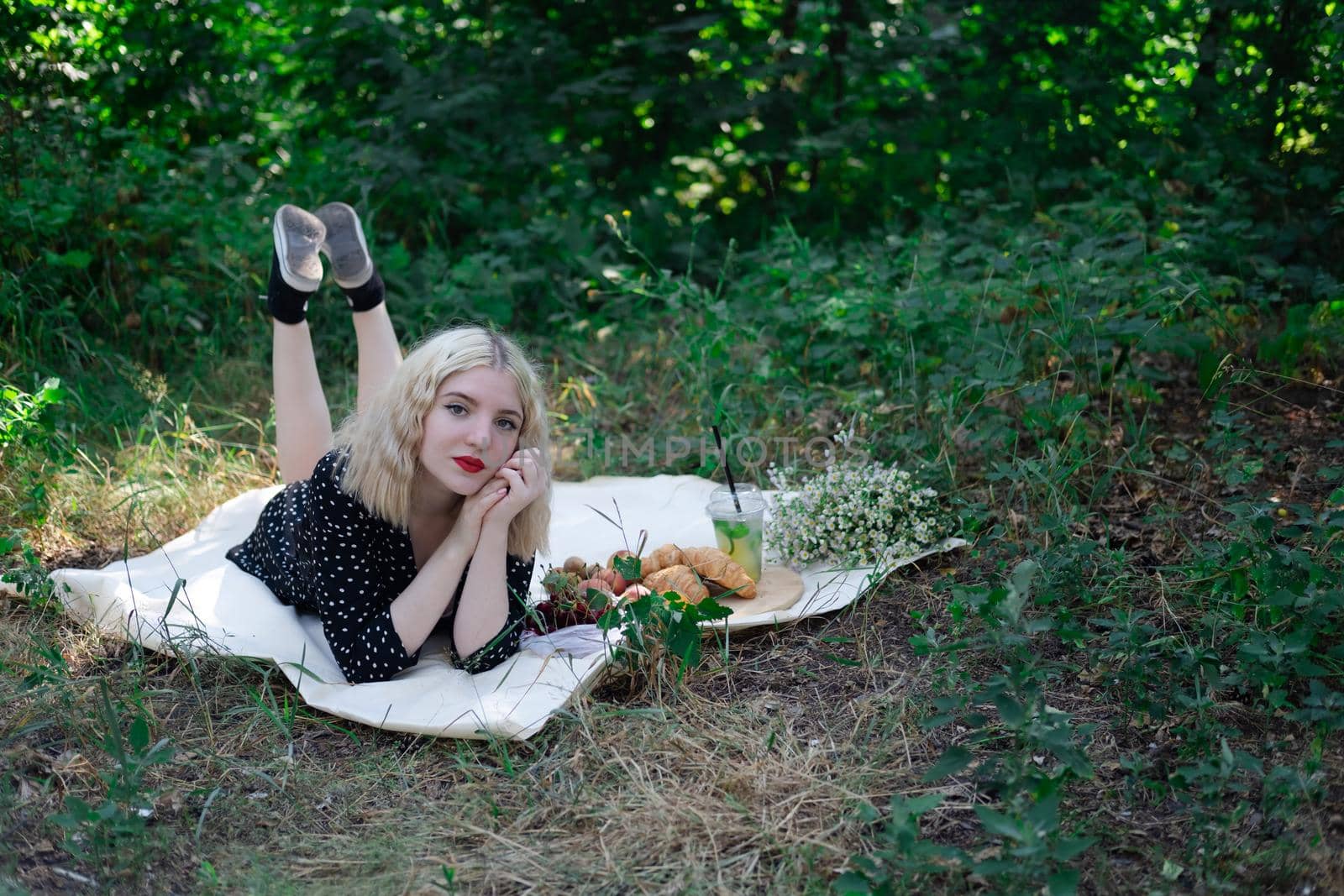 charming blonde young woman on a picnic on plaid in park with tasty snacks. Lemonade, fruits and croissants. summertime, rest, relax, enjoy. freedom.