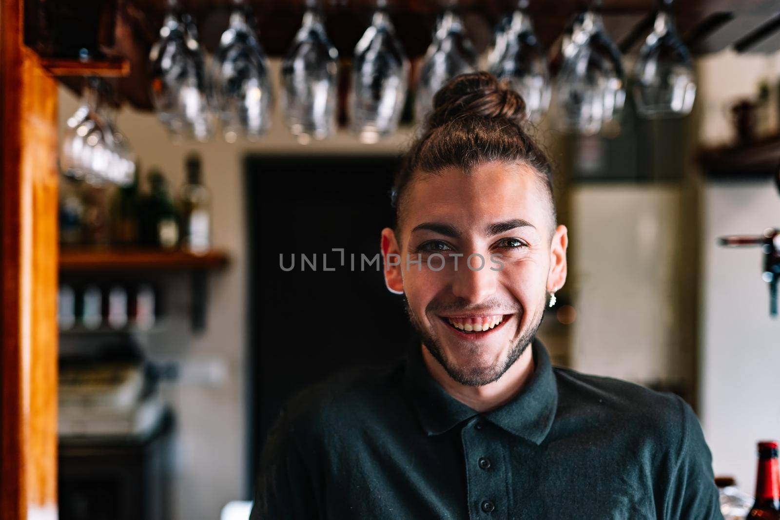 Portrait of a young, smiling, experienced, focused and hard-working waiter, dressed in company uniform, black polo shirt, standing in his small business. Looking at the camera. Warm atmosphere and dim lighting. horizontal Glasses hanging in the background over the counter.