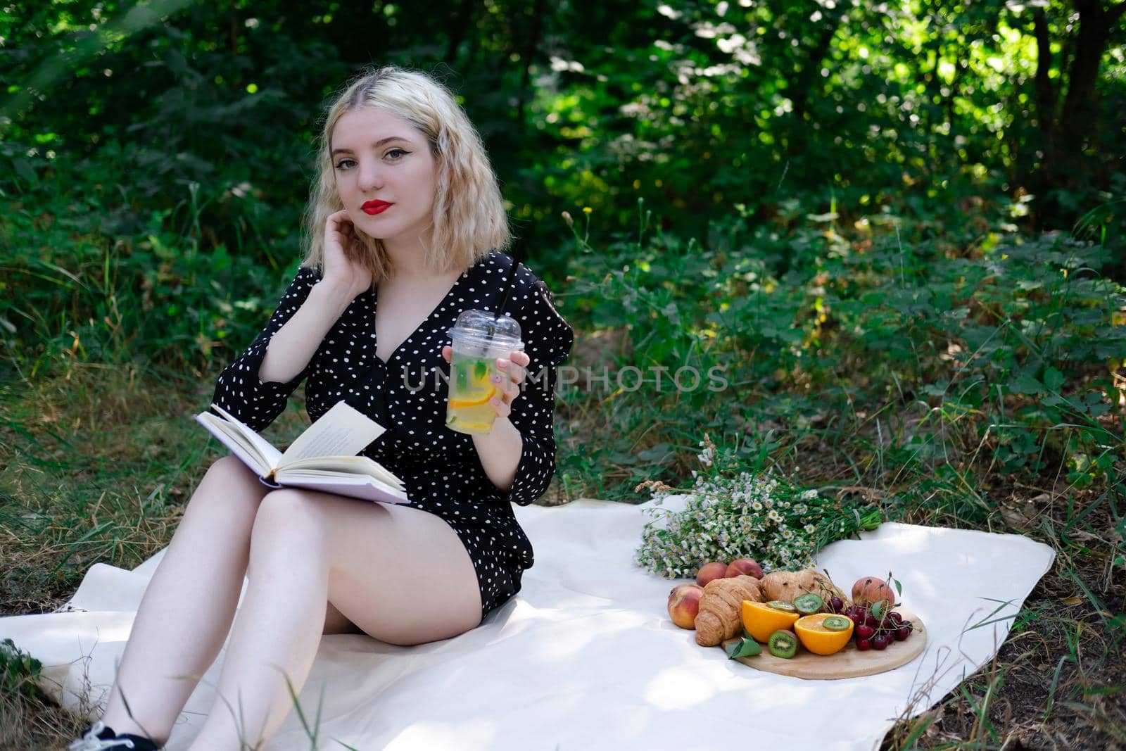 portrait of young woman on a picnic on plaid in park reading a book with tasty snacks.