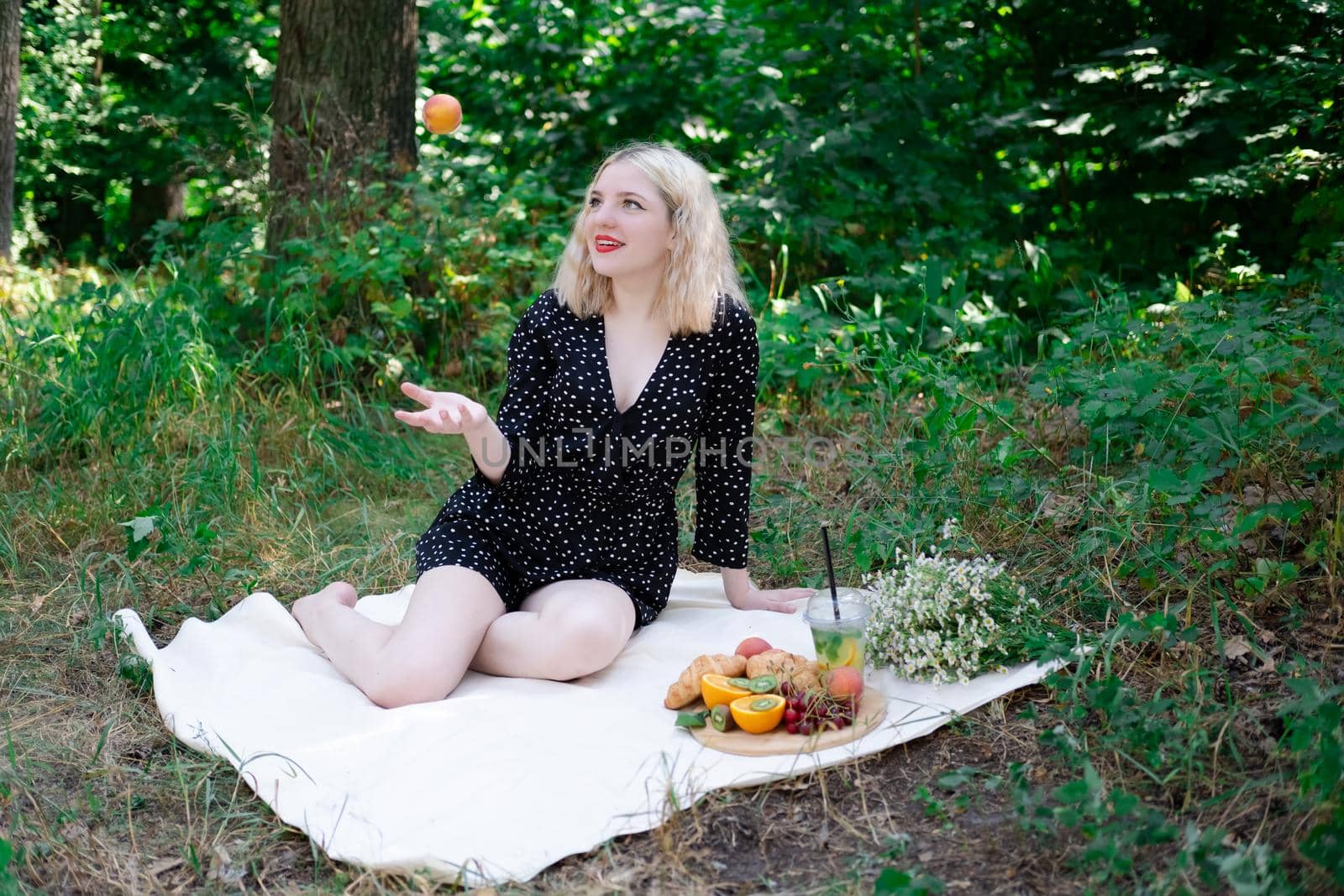 charming blonde young woman on a picnic on plaid in park with tasty snacks. Lemonade, fruits and croissants. summertime, rest, relax, enjoy. freedom.