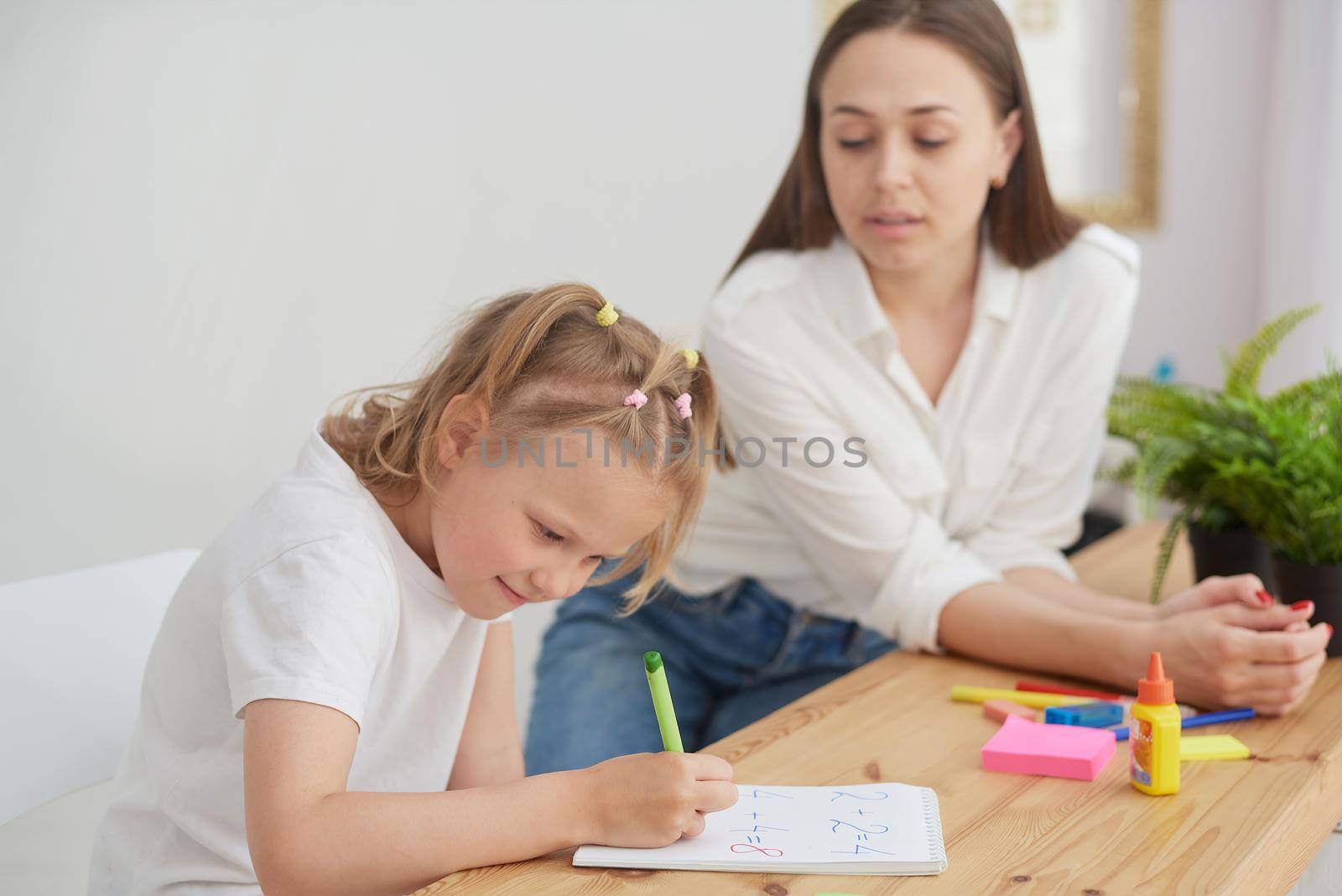 Tutor and child doing homework at kitchen. Mom and daughter are trying to solve the task. They are in a good mood and smile.