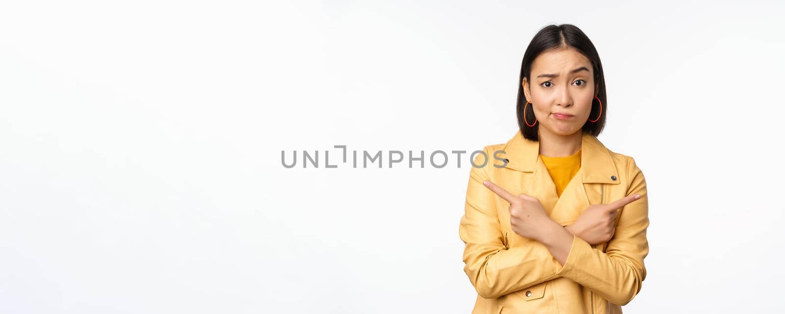 Image of indecisive asian girl, pointing fingers sideways, pointing left and right, choosing variant, deciding, standing over white background. Copy space