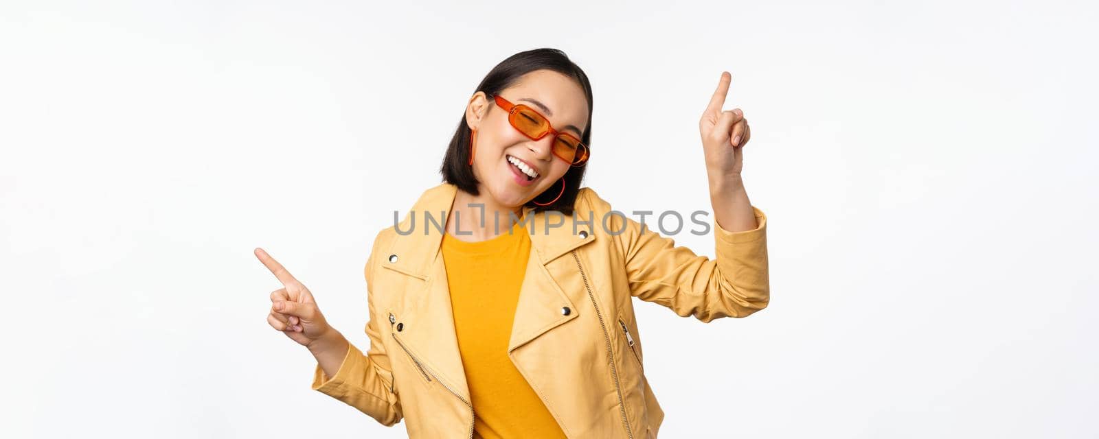 Smiling asian brunette woman in sunglasses, pointing fingers sideways, left and right, showing variants, laughing and dancing, wearing sunglasses, white background.