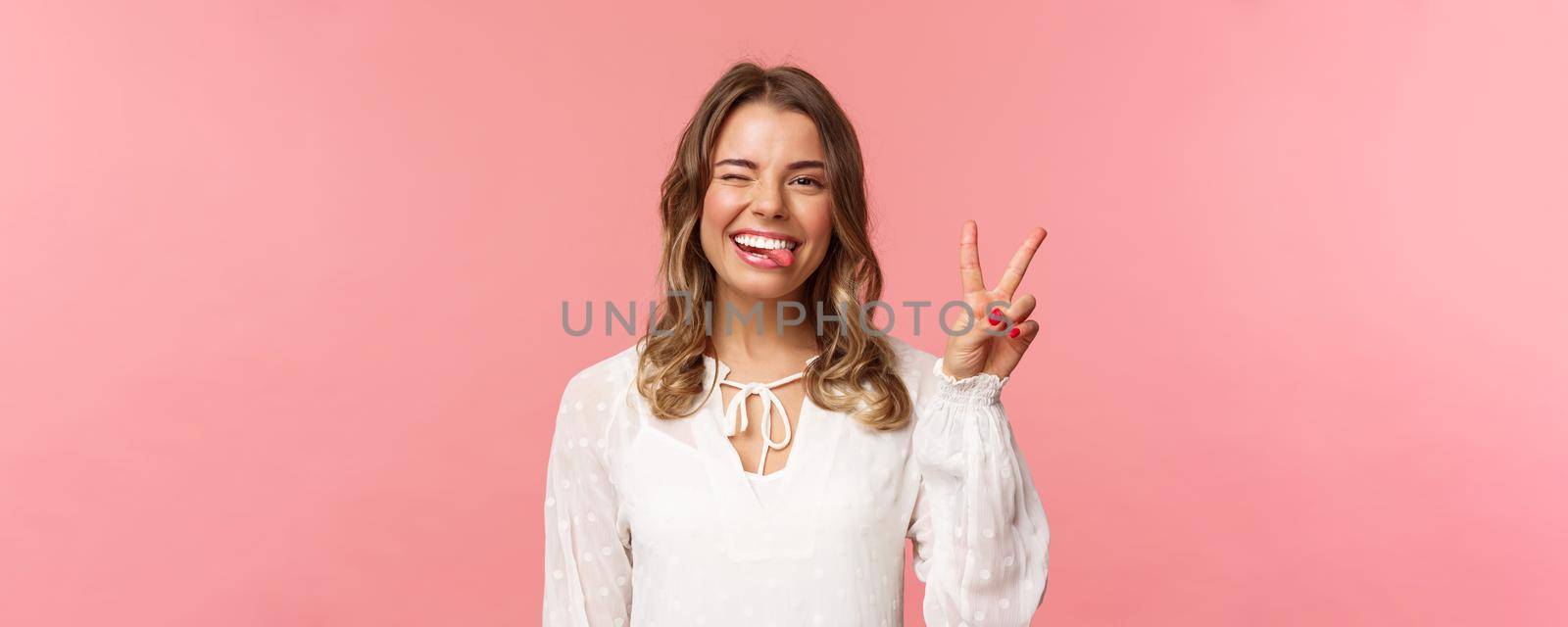 Close-up portrait of friendly joyful blond girl in white dress, sending positive vibes, smiling pleased and wink, stick tongue showing peace sign, looking kawaii over pink background.