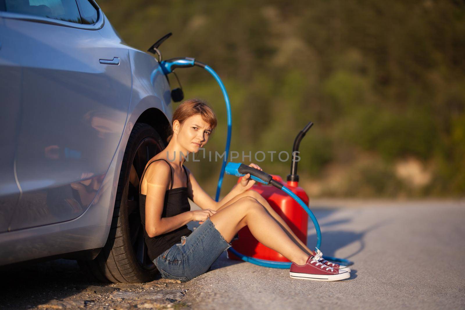 Sad girl sitting on the ground next to electric car. Holding charging cable and red gassoline canister.