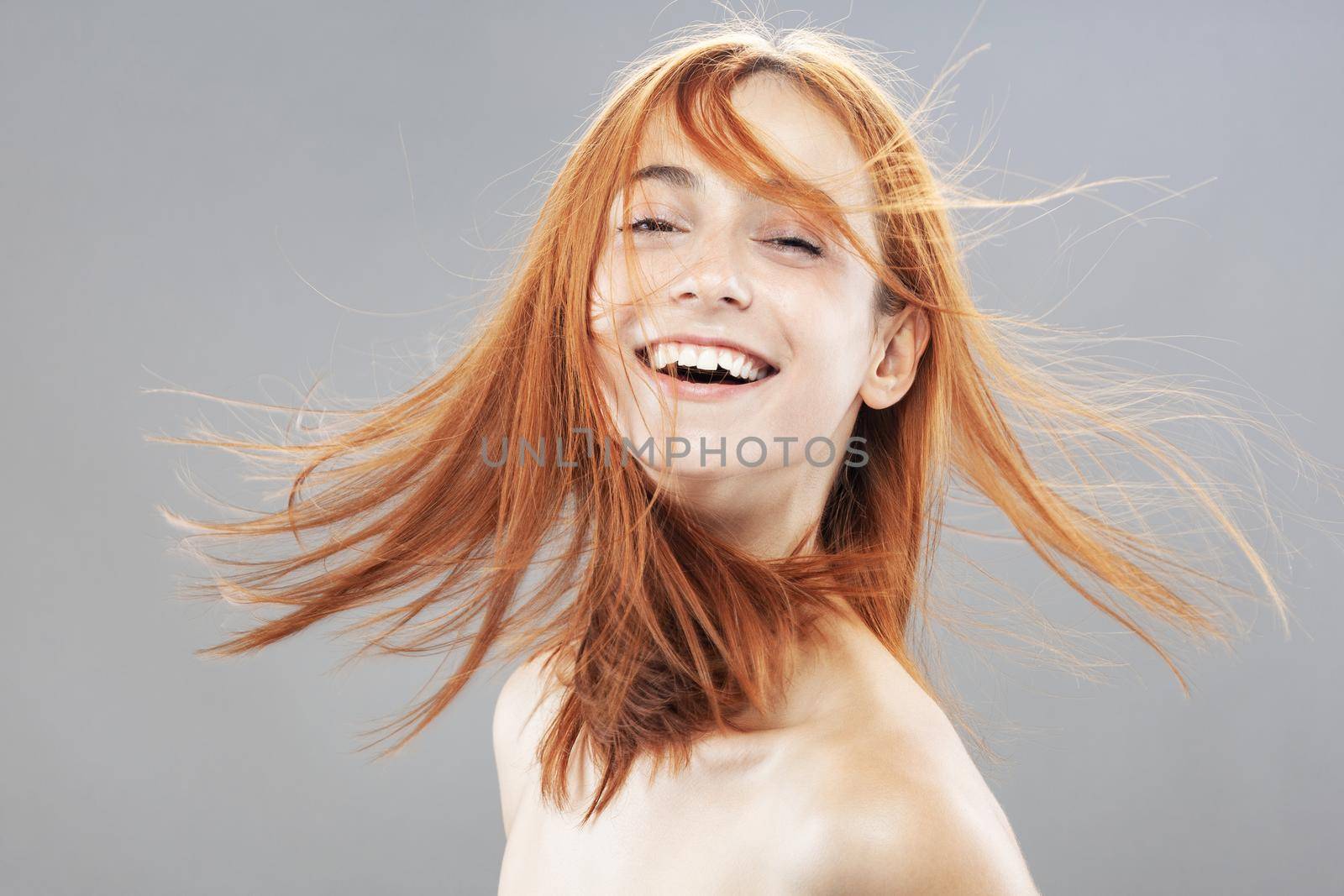 Beautiful dark burnt orange windy hair girl smiling. Studio portrait with happy face expression against gray background... by kokimk