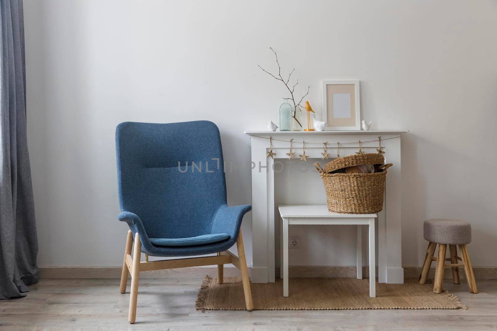 Fragment of the interior of a cozy house. A gray armchair by a white fireplace. On the fireplace is a glass bottle with a branch, figurines of birds and a photo frame. Place for text by elenarostunova