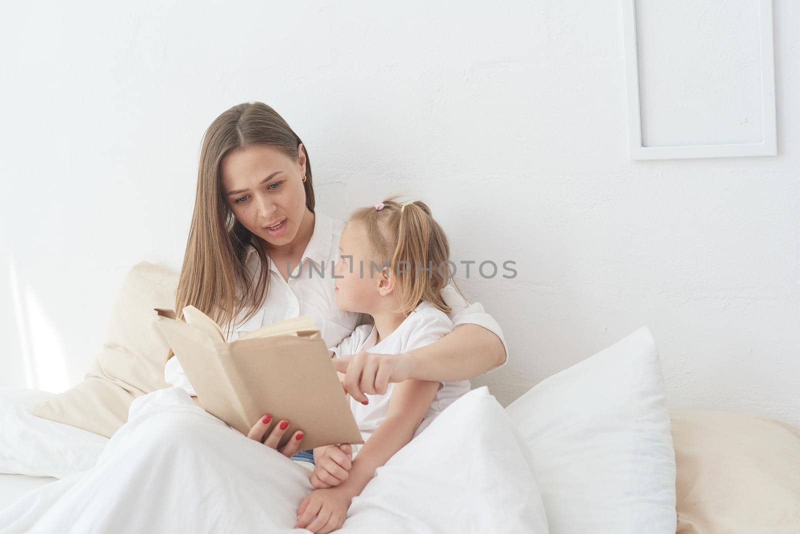 Close-up mom and little daughter are resting in a cozy bed, reading a funny interesting children's book together by etonastenka