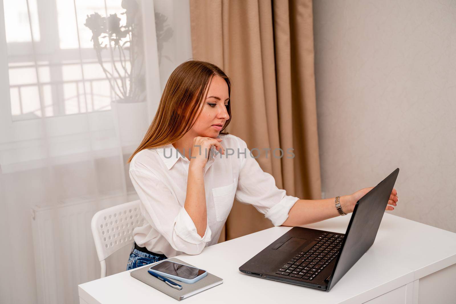 European professional woman is sitting with a laptop at a table in a home office, a positive woman is studying while working on a PC. She is wearing a beige jacket and jeans and is on the phone