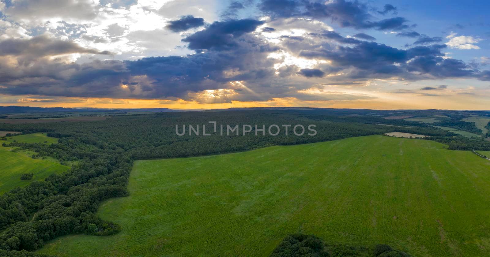 Amazing panoramic view of cloudy sky with sun rays over green field and forest by EdVal