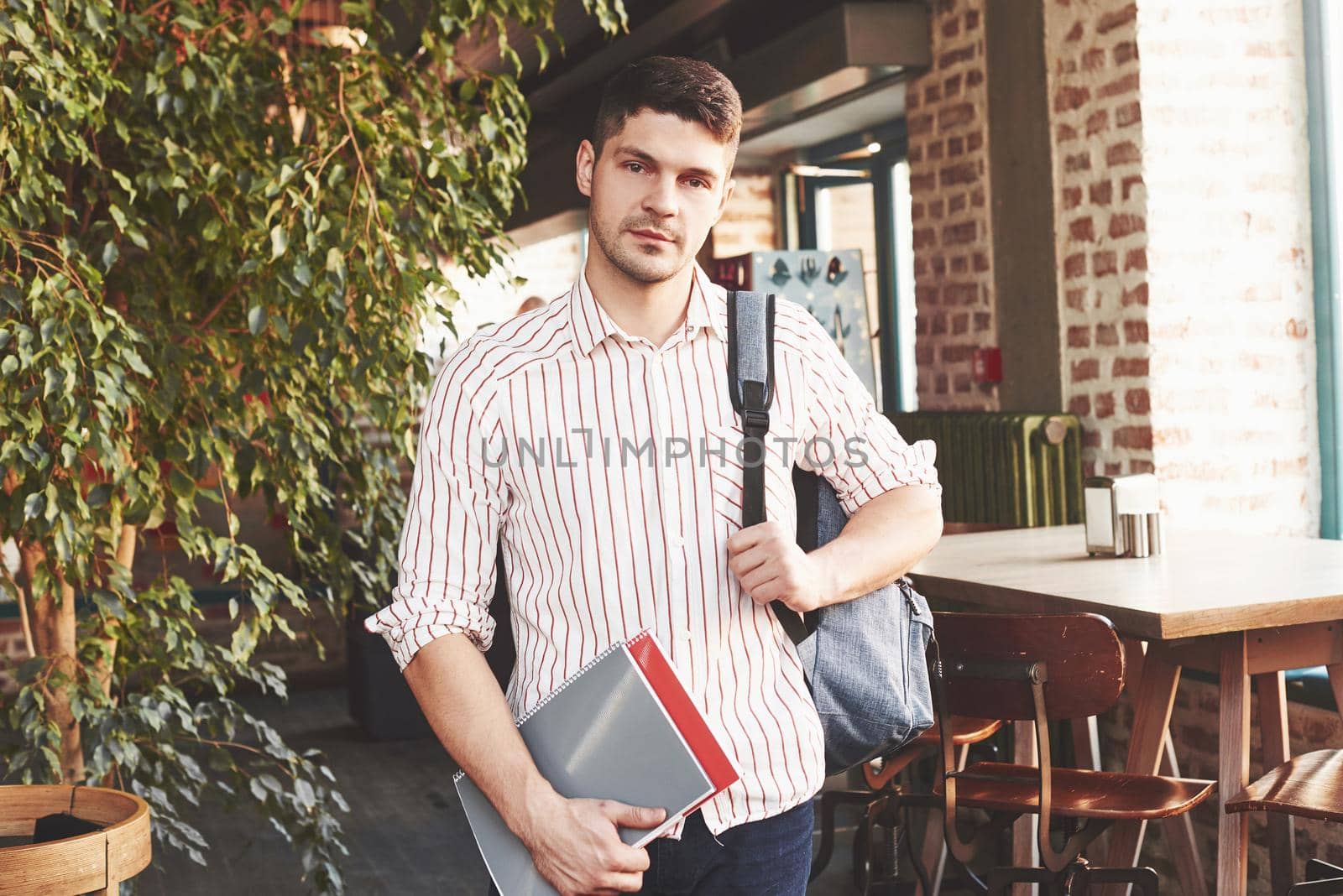 At the break time. Man in casual clothes and backpack stands indoors and holds notepads.