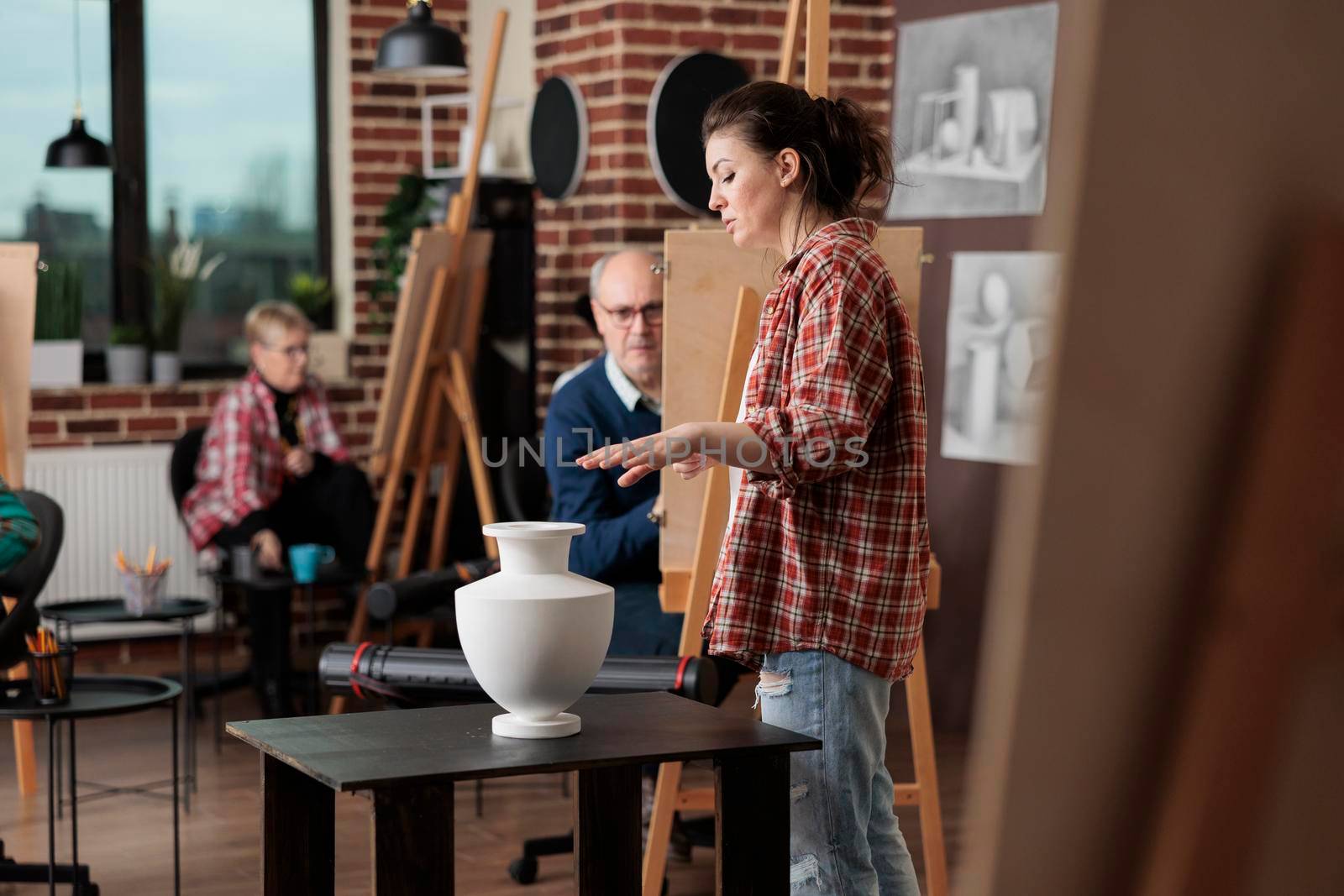 Young painter teacher supervising art class explaining graphic technique to diverse students in creativity studio. Multiethnic people sketching vase model on painting canvas learning artistic skill